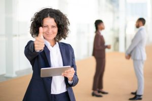 Happy businesswoman recommending new business app on tablet. Business woman with tablet standing in hallway, smiling at camera and showing thumb up. Digital technology or like gesture concept